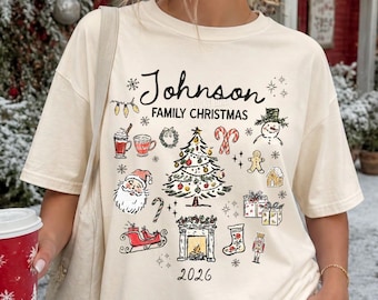 May include: An ivory t-shirt featuring a graphic print with the text Johnson Family Christmas 2026. The design includes illustrations of a Christmas tree, Santa Claus, a snowman, a fireplace, a sleigh, gifts, a stocking, and holiday treats.