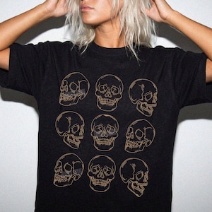 May include: A black short-sleeve t-shirt featuring a grid of nine gold-colored line art skull illustrations. The skulls are arranged in three rows of three, displaying various angles of the human cranium, including front-facing and profile views.