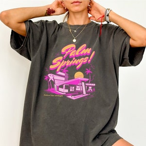 May include: An oversized t-shirt in dark grey with a pink and orange graphic print of a retro motel with palm trees and the text "Palm Springs!"