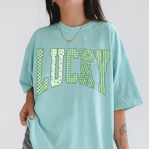 May include: A light blue t-shirt with the word "LUCKY" in green, checkered, and striped patterns. The shirt is a relaxed fit and has short sleeves. The text "CHALKY MINT" is at the bottom.