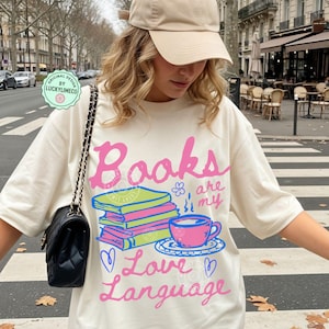以下が含まれることがあります： 「Books are my Love Language」のピンクと青の文字がプリントされたアイボリー色のTシャツ。本の山、ティーカップ、ハートのグラフィック。ベージュのベースボールキャップと黒いハンドバッグがコーディネートを完成させています。
