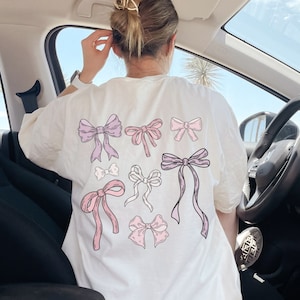 May include: White t-shirt with a pink and purple bow design. The shirt has the word "WHITE" printed on the bottom right corner.