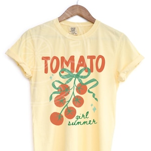 May include: A light yellow t-shirt with the word "TOMATO" in red block letters above a green bow and a cluster of red tomatoes. The words "girl summer" are in green script below the tomatoes. The shirt is hanging on a wooden hanger.