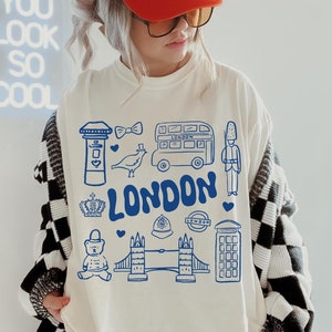 May include: A white sweatshirt with a blue graphic design of London landmarks, including a double-decker bus, a red telephone booth, Big Ben, and the words "LONDON".