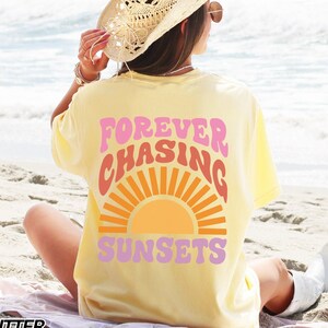 Może przedstawiać: Żółta koszulka z napisem "FOREVER CHASING SUNSETS" w różowych i pomarańczowych literach ze stylizowanym pomarańczowym i żółtym słońcem.