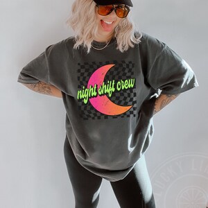 May include: A black t-shirt with a graphic of a crescent moon with a pink and orange gradient on a black and white checkered background. The text "night shift crew" is written in green on the moon.
