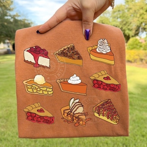 May include: A rust-colored t-shirt featuring a variety of pie slices in a cartoon style. The pies include cherry, pecan, pumpkin, and apple, some topped with whipped cream. The shirt is held up by a hand.