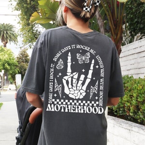 May include: Pepper-colored t-shirt with a white graphic on the back. The graphic includes a skeleton hand making a rock-and-roll sign, butterflies, lightning bolts, and the word "MOTHERHOOD". The text reads "SOME DAYS I ROCK IT. SOME DAYS IT ROCKS ME. EITHER WAY WE'RE ROCKIN".