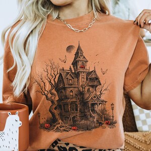 Może przedstawiać: Pomarańczowy t-shirt z czarno-białą ilustracją przerażającego nawiedzonego domu z pełnią księżyca i latającymi wok&oacute;ł nietoperzami.