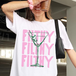 Pode incluir: Camiseta branca oversized com um gr&aacute;fico rosa de um copo de martini com azeitonas e o texto "Filthy Filthy Filthy".