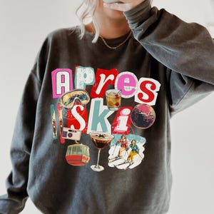 May include: Dark gray sweatshirt with the text "Apres Ski" in colorful block letters. The graphic includes images of ski goggles, a cocktail, a disco ball, a camera, a cable car, a martini, and skiers. The sweatshirt is a relaxed fit.