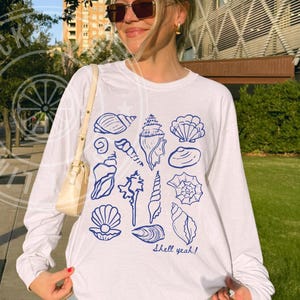 May include: White long-sleeve shirt with a blue seashell graphic and the text "shell yeah!". The shirt is made of a soft material and is perfect for casual wear. The design features various seashell illustrations.