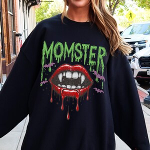 May include: Black sweatshirt with a graphic print of a vampire mouth with fangs and dripping blood. The text "MOMSTER" is printed above the mouth in green.
