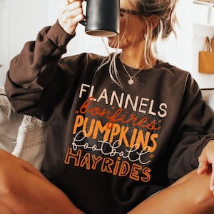 May include: A dark chocolate-coloured sweatshirt featuring the words "Flannels, Bonfires, Pumpkins, Football, Hayrides" in white, orange, and brown lettering. The person is holding a black mug. The sweatshirt is paired with fuzzy slippers.