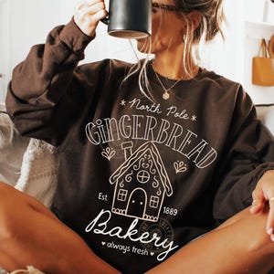 Może przedstawiać: Ciemnobrązowa bluza z białym nadrukiem w postaci domku z piernika i napisem "North Pole Gingerbread Bakery". Bluza ma okrągły dekolt i długie rękawy. Czarny kubek jest trzymany przed twarzą.