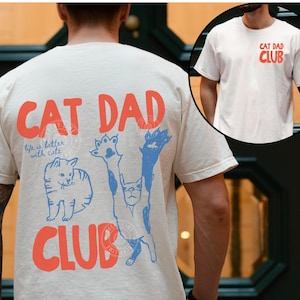 May include: Ivory-colored t-shirt with the text "CAT DAD CLUB" in coral. The back of the shirt features blue cat illustrations and the phrase "life is better with cats." A smaller image shows the front of the shirt with the same text.
