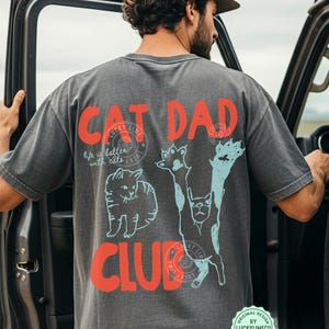May include: Dark gray t-shirt with the words "CAT DAD CLUB" in red, and cat illustrations in light blue and white. The shirt also has the text "life is better with cats". The shirt is worn by a person standing next to a vehicle.