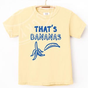Op de afbeelding: Een geel T-shirt met een blauwe afbeelding van een banaan en de tekst "THAT'S BANANAS".