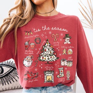 May include: Crimson-colored long-sleeve sweatshirt with a festive graphic. The design includes a Christmas tree, Santa Claus, and the text "'tis the season." Other illustrations include Christmas lights, candy canes, and a fireplace.