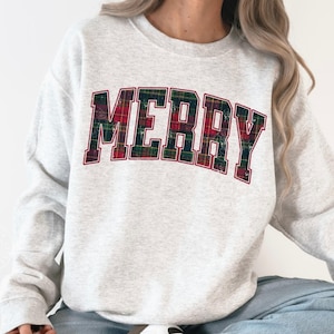Könnte beinhalten: Hellgraues Sweatshirt mit dem Wort "MERRY" in einem Schottenkaro-Design auf der Brust. Das Sweatshirt wird mit hellblauen Jeans und weißen Turnschuhen kombiniert. Das Wort "ASH" ist in der unteren rechten Ecke aufgedruckt.