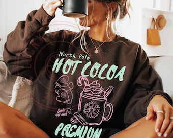 May include: A dark chocolate-coloured sweatshirt featuring the text "North Pole HOT COCOA" in a mint green font. The graphic includes a gingerbread man and a mug of hot cocoa. The sweatshirt is part of a Christmas-themed collection.