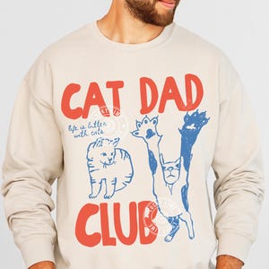 Puede incluir: Sudadera color arena con el texto "CAT DAD CLUB" en rojo. El diseño incluye ilustraciones de gatos azules y blancos y la frase "life is better with cats". Una pegatina redonda "ORIGINAL DESIGNER" está en la parte inferior izquierda.