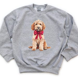 May include: Sport grey sweatshirt with a seated, light brown dog wearing a red bow. The dog has curly fur and is centered on the front. The text "SPORT GREY" is printed in black at the bottom right corner of the sweatshirt.