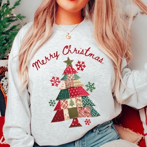 May include: A light gray sweatshirt with the words "Merry Christmas" in red script above a patchwork Christmas tree design. The tree is made of red, green, and tan fabric patches, with red snowflake accents.