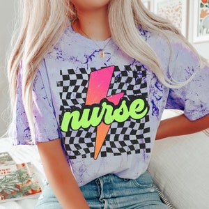 May include: A purple tie-dye t-shirt with a black and white chequered pattern and a lightning bolt graphic. The lightning bolt is pink, orange, and yellow. The word "nurse" is written in green letters on the lightning bolt.