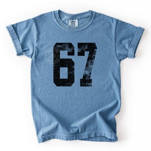 May include: A light blue t-shirt with the number 67 printed in black. The shirt has rolled-up sleeves and a crew neck. The text "BLUE JEAN" is at the bottom right corner.