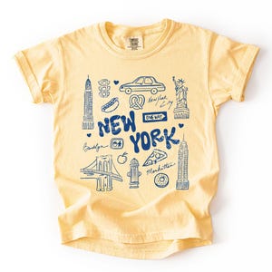 May include: Butter yellow t-shirt with a blue graphic of New York City landmarks, including the Statue of Liberty, the Empire State Building, and the Brooklyn Bridge. The words "New York" are prominently displayed.