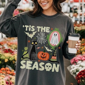 Może przedstawiać: Ciemnoszara bluza z długim rękawem z nadrukiem graficznym zawierającym napis 'TIS THE SEASON, czarnego kota, dynię, ducha w ramce, kapelusz czarownicy, miotłę, nietoperze oraz pajęczynę.