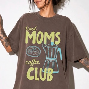 May include: A dark brown short-sleeve t-shirt featuring the text "tired MOMS coffee CLUB" in light green. The graphic includes a light blue outline of a moka pot and a small pastry on a plate. The shirt has a crew neckline and a loose fit.