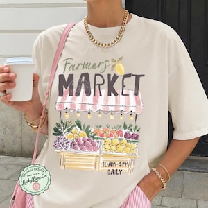 May include: Ivory-colored t-shirt with a Farmers Market graphic. The design features a market stall with fruits and vegetables, the text "Farmers Market", and the hours "10AM-5PM Daily". The shirt is paired with pink striped shorts.