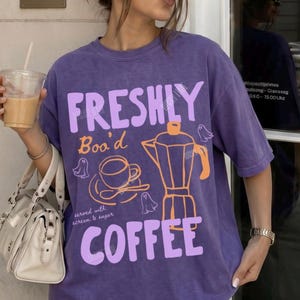 May include: A grape-coloured short-sleeve t-shirt featuring the text "FRESHLY Boo'd COFFEE" in light purple. The graphic includes an orange moka pot, a coffee cup, and small ghost illustrations. The shirt has a relaxed fit and a crew neckline.