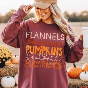 May include: A long-sleeve maroon sweatshirt featuring the text FLANNELS, bonfires, PUMPKINS, football, and HAYRIDES in a mix of block and script fonts. The garment has a crew neckline and a relaxed fit, with the text printed in orange and white.
