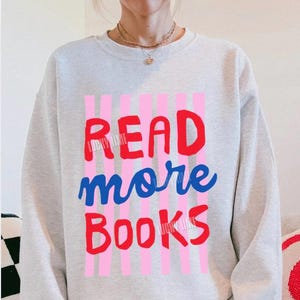 Op de afbeelding: Een lichtgrijs sweatshirt met de tekst "READ more BOOKS" in rood en blauw, tegen roze verticale strepen. Het sweatshirt maakt deel uit van een casual outfit, gecombineerd met roze en witte geruite shorts.