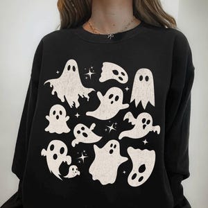 May include: Black sweatshirt with a pattern of white ghost illustrations and star accents. The word "BLACK" is printed at the bottom. A round sticker with the text "ORIGINAL DESIGN ONLY SOLD BY Lucky Lime Co" is visible.