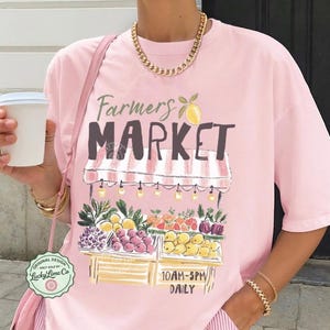 May include: A light pink t-shirt with a "Farmers Market" graphic featuring a fruit stand with various fruits and vegetables. The shirt also has the text "10AM-5PM DAILY". The shirt is part of the "Blossom" collection.