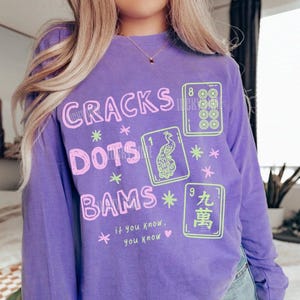 Puede incluir: Una camiseta violeta de manga larga con el texto CRACKS, DOTS y BAMS en rosa, junto a tres ilustraciones de fichas de Mahjong en verde claro. La camiseta también incluye el texto if you know, you know en letras rosas pequeñas con un corazón.