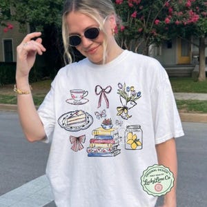 May include: White t-shirt with a charming illustration of a teacup, cake, books, flowers, and butterflies. The shirt has the word "WHITE" at the bottom left and a circular logo that says "Lucky Lime Co."
