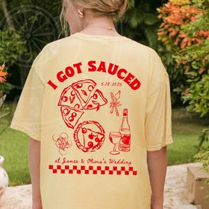 May include: A yellow t-shirt with the text "I got Sauced 5.14.2025 at James & Olivia's Wedding" in red. The shirt features a graphic of pizza slices and a glass of wine.