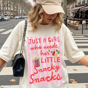 Puede incluir: Camiseta color marfil con el texto "JUST A GIRL who needs her LITTLE Snacky Snacks" en rosa y rojo. El diseño incluye una cubeta de palomitas de maíz e ilustraciones de caramelos. La persona lleva una gorra beige y un bolso negro.