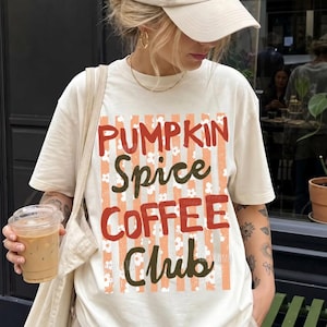 Pode incluir: Uma t-shirt de manga curta na cor creme com uma estampa gráfica de listras verticais laranja e flores brancas. O texto PUMPKIN SPICE COFFEE CLUB está escrito em letras vermelhas e verde-escuras. A t-shirt tem gola redonda e um corte descontraído.