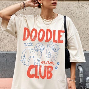 Puede incluir: Camiseta color marfil con las palabras "DOODLE MOM CLUB" en rojo. La camiseta presenta ilustraciones de perros doodle azules y la frase "life is better with doodles". La camiseta es parte del dise&ntilde;o original de Lucky Lime Co.