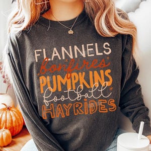 May include: A charcoal gray long-sleeve crewneck sweatshirt featuring the text FLANNELS, bonfires, PUMPKINS, football, and HAYRIDES in various orange, cream, and brown fonts. The shirt has a relaxed fit and a vintage-washed appearance.