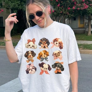 May include: A white t-shirt with a graphic of nine dogs wearing bows. The dogs are all different breeds and have different colored bows. The shirt is being worn by a person with brown hair and sunglasses.