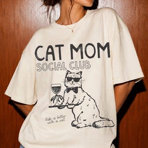 K&ouml;nnte beinhalten: Ein elfenbeinfarbenes Kurzarm-T-Shirt mit dem schwarzen Schriftzug CAT MOM SOCIAL CLUB. Unter dem Text befindet sich eine Grafik einer Katze mit Sonnenbrille und Fliege, die ein Weinglas auf einem Tablett h&auml;lt. Darunter steht der Text Life is better with a cat.