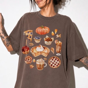 May include: An espresso-colored short-sleeve t-shirt featuring a central graphic print of various autumn-themed treats, including a pumpkin, a slice of pie, a caramel apple, a muffin, a latte, and a lattice-crust pie, all rendered in watercolor style.