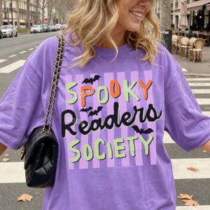 May include: A violet-colored t-shirt with the text "Spooky Readers Society" in orange, green, and black, with bat graphics. The shirt has a striped background and is a casual, oversized fit.